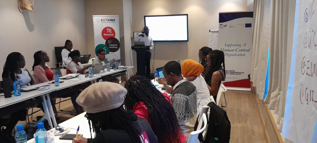 A group of women are sitting in a U-shaped formation at a conference table. Meshack Masibo is tanding in at the front and making a presentation.