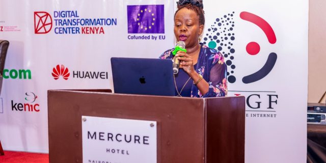 Grace Githaiga is holding a microphone and standing behind a podium labelled Mercure Hotel.