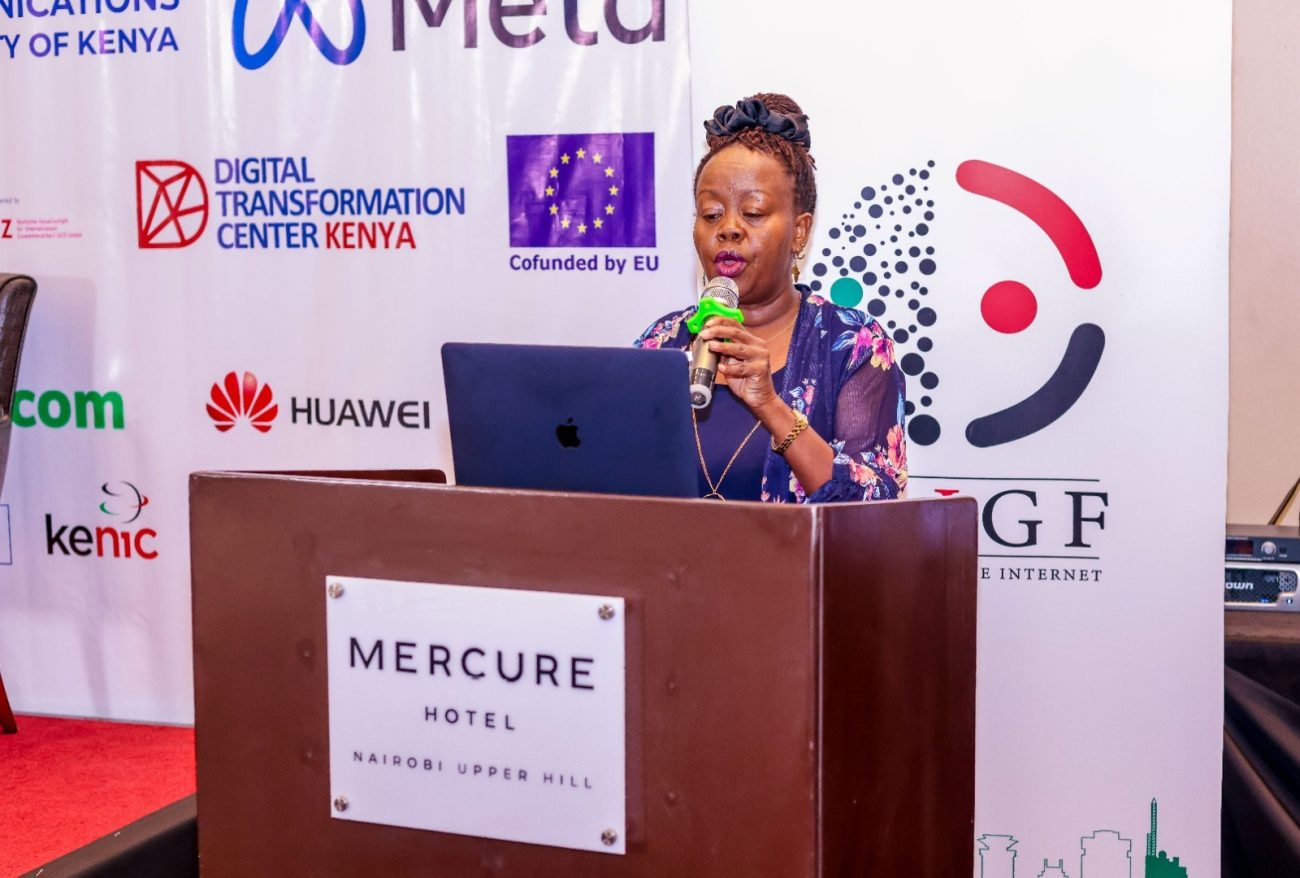 Grace Githaiga is holding a microphone and standing behind a podium labelled Mercure Hotel.