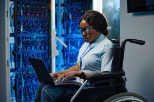 African female IT engineer with disability sitting in wheelchair and typing data on laptop during her work in data center