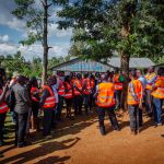 A group of men and women in orange vests are standing under a tree listening to a man in orange vest and green t-shirt