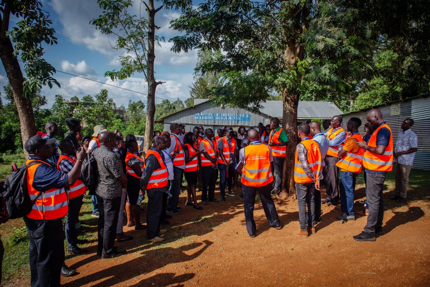 A group of men and women in orange vests are standing under a tree listening to a man in orange vest and green t-shirt